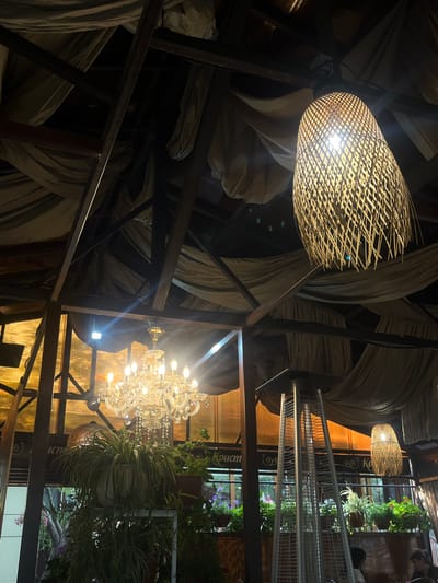 The image depicts the interior of a dimly lit establishment, possibly a restaurant or cafe. Dominating the foreground is a large, woven basket-style lampshade with a bright bulb inside, casting a warm glow. Behind it, a dramatic chandelier with multiple crystal elements and lit bulbs hangs, reflecting light. A metal structure, possibly shelving or part of the building's framework, is visible, interspersed with flowing, draped fabric in shades of grey and beige.
In the background, there are glimpses of greenery, suggesting the presence of potted plants or a garden area. A metal railing or shelf unit is also visible, holding more plants. Faint outlines of people can be seen in the distance, indicating that the establishment is occupied. The overall ambiance is one of subdued lighting and an eclectic decor, with a mix of natural materials and ornate fixtures. The time of day appears to be evening or night, given the artificial lighting. There is no indication of weather conditions.
Visible text in the image includes "Константинополь" written on a decorative element behind the chandelier. The location is identified as Novy Urengoy, Russia.
