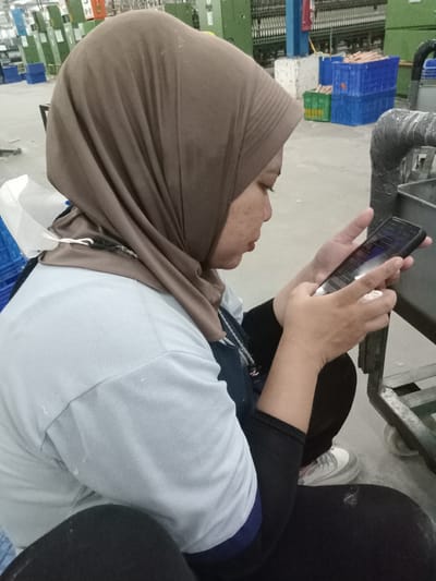 A woman wearing a light brown hijab and a light blue t-shirt with a dark blue collar is seated on the floor, looking at a smartphone. She is also wearing black leggings and white sneakers with gray accents. A white surgical mask is partially visible near her neck.
The background appears to be an industrial setting, potentially a factory, with green and blue machinery and storage bins. The location is Sukomoro, Indonesia.