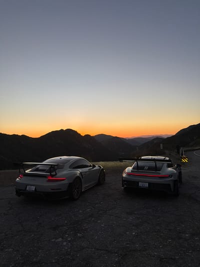 The image displays two grey Porsche sports cars, a 911 GT2 RS (left) and a 911 GT3 (right), parked on dark, cracked asphalt pavement during dusk. The vehicles are oriented with their rear three-quarters facing the viewer; both have illuminated red taillights. The left vehicle's license plate reads "TRUST," while the right vehicle's plate reads "FUNDED." The setting is a mountainous, winding road near Altadena, United States. Multiple dark mountain ranges are silhouetted against a gradient sky, transitioning from a bright orange-yellow at the horizon to a pale blue overhead. A yellow and black striped hazard marker is visible on the roadside to the far right. No individuals are present in the scene.
