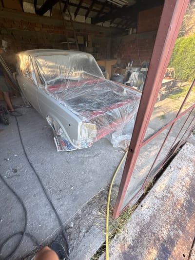 In Radinovo, Bulgaria, a car, partially covered in clear plastic sheeting, sits in a garage. The car's exterior appears to be undergoing preparation for painting, with some parts showing a red primer. The garage environment is cluttered, with tools, a ladder, and various other items visible in the background. The floor of the garage is concrete, with some debris scattered around. The scene suggests an ongoing automotive restoration or repair project.