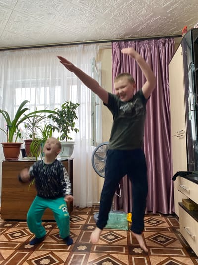 A photo depicts two boys indoors. One boy, in a green t-shirt and dark pants, is leaping in the air. The other boy, wearing a black and white patterned shirt and green pants, appears to be standing on the floor. In the background, there are sheer curtains, a potted plant, a fan, a curtain, a TV, and a tiled floor. The photo was taken in a place that has been identified as Bely Klyuch, Russia.