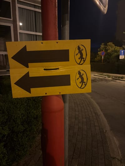 The image depicts a street scene at night in Strombeek-Bever, Belgium. Two yellow directional signs, each with a black arrow pointing left and a black silhouette of a gecko within a circle, are attached to a red pole. The signs are mounted one above the other. To the left of the pole is a building with windows covered by blinds. To the right of the pole, a paved sidewalk runs alongside a road. In the background, there are trees and street signs, including a blue sign with a white arrow pointing upwards. The overall atmosphere is dark, indicating it is nighttime.