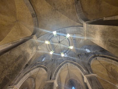 This is a wide-angle, low-angle shot looking up at the interior ceiling of the Crusader church in Abu Gosh, Israel. The stone arches and domed ceiling are illuminated by a large, circular chandelier with eight rectangular, frosted glass light fixtures. The stone work is aged and textured, with visible mortar lines and variations in color from pale yellow to grey. Several small, arched windows with decorative grilles are visible in the upper parts of the walls. Sunlight streams through some of the windows, creating bright lens flares and illuminating the dust motes in the air. The overall impression is one of ancient architecture and atmospheric lighting.