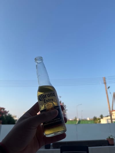 A hand is holding a Corona Extra beer bottle against a clear blue sky with visible power lines. The bottle has a lime wedge in it and the label is clearly visible. In the background, there is a blurry view of buildings and a street light, consistent with an urban or suburban environment. The lighting suggests it is daytime, likely late afternoon or early morning due to the warm tones and shadows. The presence of power lines and buildings indicates this is likely an outdoor setting in an inhabited area. The overall scene evokes a casual, relaxed atmosphere, possibly on a balcony or patio. This scene could be located in Paralimni, Cyprus, given the characteristic clear blue skies and urban infrastructure.
