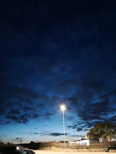 This image depicts an outdoor scene in Montuïri, Spain, at twilight or night, illuminated primarily by artificial light. The upper two-thirds of the frame are dominated by a deep blue sky with widespread, dark, low-lying clouds. A single distinct, bright white point, possibly a star or planet, is visible in the upper central-left portion of the sky.

A tall, slender, white streetlamp stands centrally, emitting a bright, warm white light that casts an intense glow directly beneath it. The ground immediately surrounding the base of the streetlamp appears to be a light-colored, unpaved surface, possibly gravel or dirt.

In the midground, a light-colored concrete or rendered wall extends horizontally across the scene, forming a barrier. Below this wall, a low, rough stone wall is visible. To the far left, a dark-colored vehicle, likely a car, is partially visible with its front hood and windshield reflecting light. To the far right, another dark-colored car is parked parallel to the low stone wall.

Behind the walls, on the right side, a building with illuminated windows or openings is visible, suggesting an active structure. Adjacent to this building, a large, dense green tree with abundant foliage is present. In the background to the left of the streetlamp, a utility pole or similar structure is discernible. No individuals are present, and no actions or interactions are depicted.