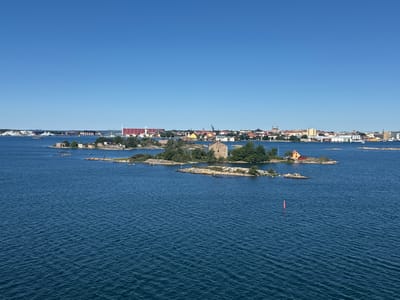 The image shows a view of the archipelago of Karlskrona, Sweden, looking towards the city. The foreground is dominated by a large expanse of dark blue, rippling water. In the middle ground, several small, rocky islands are scattered across the water. Some of these islands are covered in green vegetation, and a few have small buildings or ruins. One island features a prominent stone structure resembling a tower. Further in the distance, the cityscape of Karlskrona is visible, characterized by a collection of buildings with red roofs and some taller structures, including cranes. The sky is a clear, bright blue. Several ships are visible in the water, both near the islands and further out towards the city. A red buoy is present in the water in the right mid-ground.