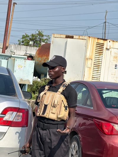 An outdoor, daytime scene in Ajah, Nigeria, features a male individual of apparent African descent, standing between two parked sedans. He is positioned centrally, facing slightly to his right (viewer's left), with a neutral expression. He wears a black polo shirt, black trousers, a black belt, and a black baseball cap with a white logo. Over his shirt, he wears a tan/beige chest rig equipped with multiple pouches. In his left hand, he holds a black two-way radio. His right hand is held open, palm upwards, at waist level.
To the individual's immediate left, the rear quarter and taillight of a silver sedan are visible. To his immediate right, the rear quarter and taillight of a dark red sedan are visible.
In the background, from left to right, are several structures under a clear, light blue sky traversed by overhead power lines. These include two rusted metal poles, a light blue/grey utility box, and a prominent rusted cylindrical metal tank. Behind the utility box and wall, a partial human figure is visible. Further right, behind the red sedan, is a white, heavily rusted corrugated metal structure, possibly a generator housing. Green foliage is present between and behind some of these structures.