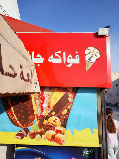 A young girl with long brown hair tied in a ponytail and wearing a light pink t-shirt and grey pants walks past a red sign advertising ice cream. The sign features white Arabic text that reads "فواكه جنان" (Fawakeh Jinan), meaning "Fruit Paradise". An image of an ice cream cone topped with chocolate and nuts is also visible on the sign. Below the red sign, a colorful advertisement displays images of ice cream bars, chocolate, nuts, and caramel candies. The scene appears to be outdoors during the day, with a clear blue sky. The setting suggests a street-side vendor or shop in a sunny climate. The girl's back is to the camera as she walks away, and her presence adds a sense of everyday life to the scene. The overall impression is one of a warm, bright day, with the vibrant advertising creating a lively atmosphere.