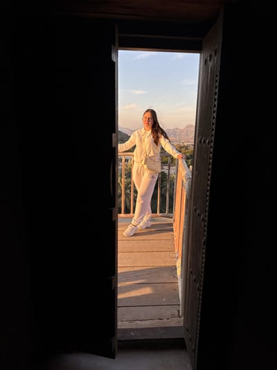 A female subject is depicted standing on a wooden balcony, viewed through a dark, open wooden doorway. She has long brown hair and is wearing a cream-colored long-sleeved top, matching sweatpants with a dark logo on the left thigh, white sneakers, and a light-colored crossbody bag. She is smiling faintly with both hands resting on a light-colored wooden railing. The balcony floor is composed of horizontal wooden planks, illuminated by warm, golden sunlight casting shadows. Beyond the balcony, a mountainous landscape with rugged peaks and a valley filled with numerous green palm trees is visible under a clear, light blue sky. This scene is situated in the city of Hatta, United Arab Emirates.
