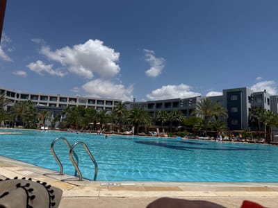 The image depicts a large outdoor swimming pool at a resort in Yasmine Hammamet, Tunisia. The pool is a vibrant blue and reflects the clear sky above, which is dotted with white, fluffy clouds. Palm trees line the edge of the pool, casting shadows on the water and surrounding area. In the background, a multi-story hotel building with a modern architectural style curves around the pool, its balconies and windows visible. Several people are seen relaxing by the pool, some sunbathing on lounge chairs, while others are in the water. A set of chrome ladders leads into the pool in the foreground. Part of a straw hat with black pom-poms is visible in the bottom left corner, suggesting the perspective of someone seated by the pool. The overall scene conveys a sense of leisure and vacation.