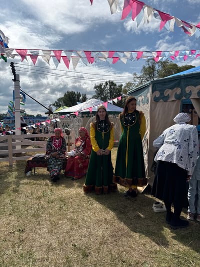 In Старое Субханкулово, Russia, a festival scene unfolds under a partly cloudy sky. A group of people are gathered outdoors near a white wooden fence and tents decorated with festive triangular flags in pink, white, and light blue. In the foreground, two young women stand dressed in traditional green and yellow attire, accented with fur trim and intricate dark beadwork on their vests. Their headwear consists of golden bands. To their left, two older women are seated on a bench covered with a patterned textile. The woman on the left wears a vibrant red headscarf and a long, colorful patterned dress, while the woman on the right has a red headscarf and a red dress, both adorned with beaded necklaces. Behind them, several other individuals are visible, including a person in a white patterned jacket and black skirt, and a child in blue jeans. The ground is covered in dry, yellowed grass, casting long shadows from the people and structures. In the background, more tents and trees can be seen, along with a distant white stork on a pole. Balloons and signage suggest a public event or fair.