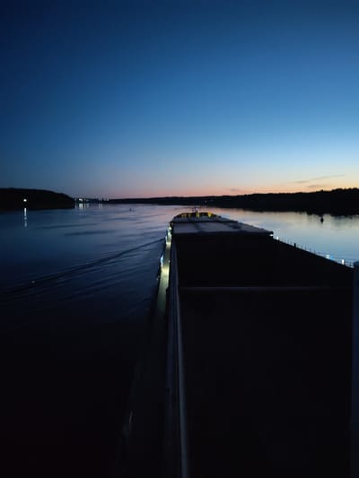 An aerial view captured at dusk or dawn shows a large cargo ship moving down a wide, calm river. The sky transitions from a deep, clear blue at the top to lighter shades of blue and a faint hint of orange near the horizon. The water reflects the sky and is disturbed by the ship's wake, creating ripples. On the left side of the river, a dark, tree-lined shore is visible, with distant lights indicating a settlement. On the right side, the foreground is dominated by the deck of the cargo ship, which is dark and appears to be carrying a load. The ship has a few lights visible along its side. The scene is set in a natural environment with a river and forestation, suggesting a location outside of a major urban center. The image does not contain any people or explicit interactions. There is no information to suggest the location is the city of Михайловский, Russia.