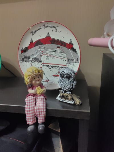 The image shows a shelf with several decorative items. On the left is a ceramic plate with a depiction of a cityscape and a boat. To the right of the plate are two figurines: a doll-like figure in a yellow hat and pink plaid pants, and a small owl figurine perched on a base. The background is a plain, textured wall. The items are arranged on a dark-colored shelf. The inscription on the plate reads "Нижний Новгород" (Nizhny Novgorod), indicating that the plate is a souvenir from that city. There is no indication of the location being Ostafievo, Russia.