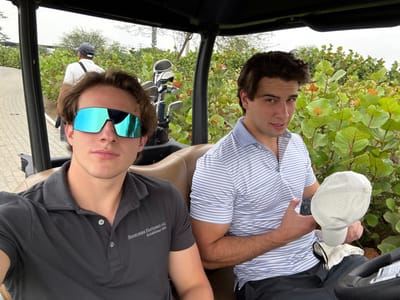 Two men are seated in a golf cart with golf clubs visible behind them. The man on the left is wearing a striped shirt and holding a white golf hat and glove. The man on the right is wearing a dark grey polo shirt with "Berkshire Hathaway Inc." printed on it and mirrored sunglasses. They appear to be on a golf course, as greenery and a paved path are visible in the background.
