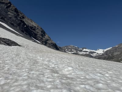 The image shows a mountain landscape with a large expanse of snow in the foreground. The snow has a textured, rippled surface. To the left, a steep, dark, rocky mountain slope rises sharply against a clear blue sky. Further in the distance, more snow-capped mountains and rocky peaks are visible, with a small, wispy cloud near the horizon. The scene appears to be a remote, natural environment, likely at a high altitude. The location is identified as Leukerbad, Switzerland. There are no people or man-made objects visible in the image.