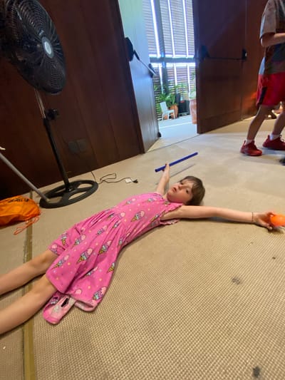 The scene appears to be indoors, likely within a residence. A young girl wearing a pink dress with an ice cream cone pattern is lying on a tan carpeted floor. She holds a blue cylindrical object in one hand and an orange ball in the other. To the left, there is a dark wooden structure, possibly a cabinet or wall. A standing fan is partially visible in the lower left corner. Another child's legs are visible in the upper left corner. An orange bag is near the fan. The location in the city of Marina Maroma, Mexico is not discernible from this image.