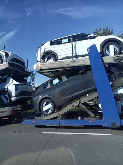 The image shows a car carrier truck transporting several vehicles. The scene is outdoors under a clear blue sky, suggesting daytime and fair weather. The truck is on a road, likely a highway, given the asphalt surface and lane markings visible in the foreground.
The subjects are primarily vehicles, which appear to be modern electric cars based on their design, particularly the distinctive wheels and body shapes. There are at least four cars visible, stacked on multiple levels of the carrier. One white car is on the top level, partially obscured. Below it, a dark gray car is angled, with its front pointing upwards and to the right. On the lower level, a white car is visible from the rear, parked on the left side of the carrier. Another white car is on the far left, partially cut off by the frame.
The activity is the transportation of these cars, likely to a dealership or distribution center. The cars are secured on the carrier, indicating a transit operation.
Notable details include the blue ramp structure of the car carrier, which is a prominent feature in the right half of the image. Some of the cars have white stickers or labels on their windows. The word "LOHR" is visible on the back of the truck on the lower left side, identifying the manufacturer of the car carrier. The overall impression is one of a routine logistics operation.
There are no people clearly visible in the image. The location context provided is Třemošnice, Czechia, which helps place the scene within a geographical area.