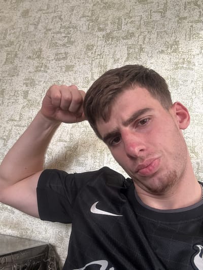 A young adult male, likely in his early twenties, with short dark brown hair and light skin, is depicted indoors. He has light brown eyes, a slight redness on his nose, and short stubble on his chin and jawline. The subject is wearing a black short-sleeved t-shirt with a white Nike swoosh logo on the left side of his chest. A silver-colored chain is partially visible around his neck. His left arm is bent at the elbow, with the hand clenched into a fist, appearing to flex the bicep. He is looking directly at the camera with a pouting expression. The background is a wall covered in light-colored wallpaper featuring a textured, abstract pattern. A dark, unidentified object is partially visible in the bottom left corner. The lighting is even. The specific location within the city of Durgena, Georgia, cannot be determined from the visual evidence provided in the image.