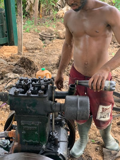 A shirtless man wearing red shorts and green rubber boots stands in front of a large, black, industrial-style engine. The man is holding a plastic bottle with some type of liquid in his right hand and an orange plastic container in his left hand. The engine is surrounded by dirt and what appears to be dried vegetation. The man appears to be working on the engine, possibly adding lubricant. The location is Egbeda, Nigeria, which is in the western part of the country. The image suggests the man is participating in a maintenance activity related to the engine.