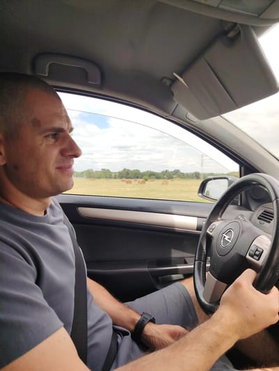 A man with a shaved head, wearing a grey t-shirt and grey shorts, is driving a car. He is looking out the passenger side window at a rural landscape with hay bales in a field. The car's interior is visible, including the steering wheel with an Opel logo and a side mirror reflecting the passing scenery. The weather appears to be overcast with some clouds in the sky. The image captures a moment of travel through a countryside setting, likely on a road near Izobil'nyi, Russia, given the presence of agricultural elements common to the region.