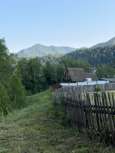 This is a vertical, eye-level, medium shot photograph taken in Anos, Russia. In the foreground, a rustic, weathered wooden fence made of vertical pickets runs diagonally from the lower right corner towards the center left of the frame. Beyond the fence, a grassy area with some recently cut vegetation is visible. In the mid-ground, a large wooden house with a corrugated roof stands behind a light-colored metal fence. The house has a balcony and appears to be elevated. To the left of the house, a dense forest of deciduous and coniferous trees extends into the background. Further in the distance, rolling, forested hills rise up, meeting a clear, pale blue sky. The overall scene depicts a rural residential area nestled within a mountainous, forested landscape.