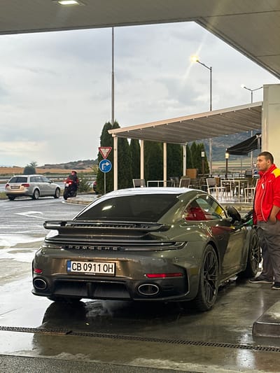 At a gas station in Drazhevo, Bulgaria, under a covered canopy, a dark grey Porsche 911 Turbo S is parked at a fuel pump. A man wearing a red and yellow jacket and grey pants stands by the car, appearing to be fueling it. The ground is wet and reflective, suggesting recent rain. In the background, other vehicles are visible, including a silver station wagon and a motorcycle with a rider. The environment appears to be an open area with trees and some distant hills under a cloudy sky. The license plate of the Porsche reads "CB 0911 OH".