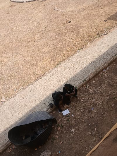 The scene is an outdoor ground-level area in Ugi Majowel, Nigeria, characterized by dry, sparse vegetation, exposed dirt, and scattered leaves. A light-colored, patterned paved walkway with a raised curb traverses the midground. Two individuals, appearing male and dressed in dark clothing, are crouched on the dirt directly adjacent to the walkway. Between them, several white sheets of paper are laid out on the ground, towards which both individuals are looking downwards. The individual on the left wears a dark long-sleeved top and dark trousers. The individual on the right wears a dark short-sleeved top and dark trousers, and has a visible chain around their neck. In the lower-left foreground, a large, dark, corrugated metal barrel lies on its side, containing visible refuse.