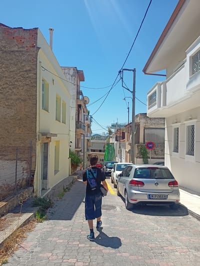 A young male walks down a narrow, sunlit street in the Municipal Unit of Anthidon, Greece. He is seen from behind, wearing a dark t-shirt with a graphic print and denim shorts, with a bag slung over his shoulder. The street is paved with interlocking stones, bordered by buildings on both sides. The building to the left is older, with exposed brick and a weathered facade, while the building to the right is a modern, white structure with decorative window grilles. Several cars are parked along the right side of the street, including a silver Volkswagen Golf with the license plate MIP-9669. The sky is clear blue. Overhead, electrical wires crisscross the sky, supported by poles. The environment appears to be a residential or commercial area.
