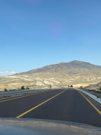 The image shows a highway under construction in a mountainous, arid landscape. The road is paved with dark asphalt and marked with bright yellow lines. Orange traffic cones are placed along the side of the road, indicating ongoing construction. In the background, a large, barren mountain range dominates the skyline under a clear blue sky. The terrain is characterized by dry, rocky soil with sparse vegetation. The overall scene suggests a remote, undeveloped area. It is not possible to determine the specific location in the city of Алғабас, Kazakhstan from the provided image.