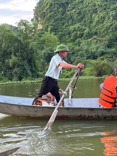 A man in a light blue polo shirt, black pants, and a green hat rows a flat-bottomed boat on a calm, green body of water. He holds a long oar with a wooden handle and a metal blade. Beside him, another person wearing an orange life vest is visible. The boat is made of metal and shows signs of wear. In the background, a lush, green mountainside covered in dense vegetation rises steeply. The water reflects the overcast sky and the surrounding foliage. The scene appears to be in a natural, possibly rural setting in Yên Sơn Ward, Vietnam, characterized by its mountainous landscape and waterways.