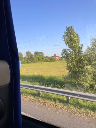 This image, taken from inside a vehicle, depicts a rural landscape under a clear blue sky. The scene is dominated by a large, grassy field that appears to have been recently mowed, with visible rows marking the cutting pattern. A line of trees borders the field, and beyond them, a red building with multiple windows can be seen. In the foreground, a metal guardrail runs along the side of what appears to be a road. The view is slightly obscured by the interior of the vehicle, with a dark blue upholstered edge and a beige headrest visible at the bottom. The time of day appears to be daytime, given the bright sunlight casting shadows. There is no visible text in the image. The overall impression is one of a peaceful, sunny day in a rural setting, possibly in Potashnia, Ukraine, given the location context.
