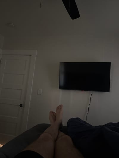 A person is lying in bed, looking at a large flat-screen television mounted on the wall. The room appears to be a bedroom, with a ceiling fan visible on the left side of the image. A white door with multiple panels is located below the television. The room is dimly lit, suggesting it might be nighttime or early morning. The person's legs and feet are visible, with one foot resting on top of the other. A dark blue blanket or duvet is covering part of the person's body. The television screen is black, indicating it may be turned off or displaying a dark image. A red light is visible on the right edge of the television, possibly an indicator light. A black cord runs from the television to the right side of the frame. A light switch is visible on the wall above the door. There are no windows visible in the frame. The overall impression is of a relaxed, domestic scene. The location is Des Moines, United States of America.