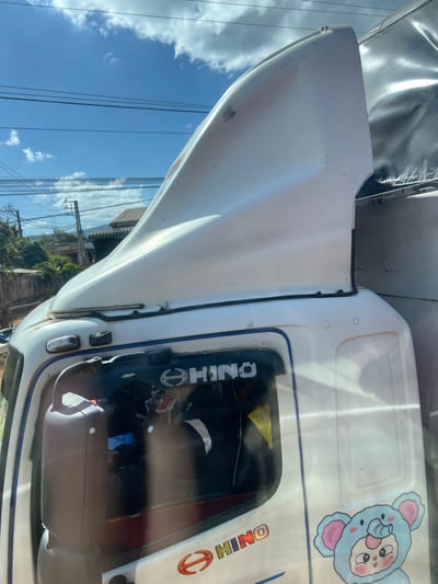 A close-up view reveals a Hino truck cab, mostly white. The front slopes downward, marked by a small indentation. The driver's side window is visible, framed by a black border showcasing a white "HINO" logo. A cartoon character sticker adorns the roof of the cab. In the reflection of the window, power lines and foliage can be seen, suggesting an outdoor setting under a partly cloudy sky.