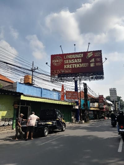 The image shows a street scene in Kecamatan Waru, Indonesia, located at coordinates -7.352 latitude and 112.729 longitude. A large billboard displays an advertisement in Indonesian for a product called "Krektek." Two men in short-sleeved shirts stand in the street near a black pickup truck parked on the side of the road. Power lines run across the scene, and other buildings can be seen in the background. In the distance, motorcycles are traveling on the road. The sky is partly cloudy.