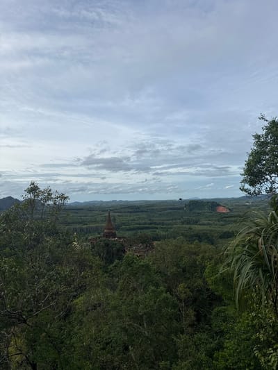 The image captures a panoramic view of a lush, verdant landscape from an elevated vantage point. In the foreground, dense green foliage from trees and other vegetation dominates the lower portion of the frame. A prominent, weathered stupa, a Buddhist religious structure, stands in the mid-ground, surrounded by more greenery and ancient ruins. The stupa is conical in shape and appears to be constructed from dark bricks. Beyond this, the landscape unfolds into rolling hills covered in an extensive forest, possibly plantations of rubber trees, stretching towards a hazy horizon. The sky above is overcast with scattered, light gray clouds, suggesting a temperate or humid climate. The scene is characteristic of the natural beauty found in areas like Ban Sadet, Thailand, known for its tropical environment and historical sites. There are no people or animals visible in the image.