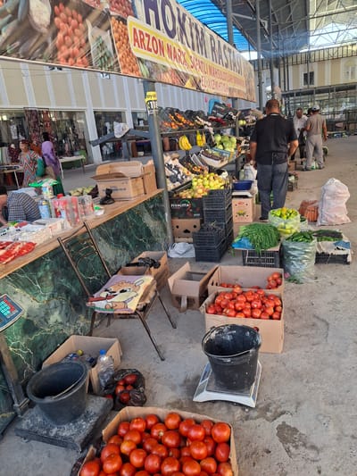The image captures an outdoor market scene in Ko`kdala, Uzbekistan. A large banner with Uzbek text, featuring images of produce, hangs overhead. The market displays a variety of fresh produce, including tomatoes in cardboard boxes, bags of carrots and peppers, bunches of green herbs, and crates of apples and bananas. Several people are present, including vendors and shoppers, interacting at the stalls. A man in a black shirt stands near a produce display, while others are seen browsing or attending to goods. The ground is concrete, and various containers, buckets, and weighing scales are visible, indicating active commerce. The overall impression is of a bustling local market.