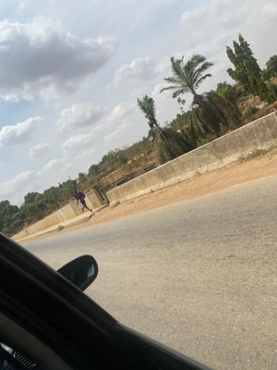 This image depicts an outdoor scene in Angwan Eggon, Nigeria, captured from the interior of a dark-colored vehicle, with a portion of the vehicle's side mirror and frame visible in the lower left quadrant. An asphalt road, light grey and textured, occupies the lower and central sections, stretching diagonally from bottom left towards the upper right. Adjacent to the road, a light brown, unpaved shoulder runs parallel to a continuous concrete barrier, approximately one meter in height, which separates the road from elevated terrain.
On the unpaved ground behind the concrete barrier, a single individual is observed walking or jogging away from the camera's perspective. The person appears to be wearing blue shorts and a dark, possibly purple, shirt. The elevated terrain behind the barrier is sparsely vegetated with dry brush and features several prominent palm trees with visible fronds, along with other taller, slender green trees. The ground on the elevated section is reddish-brown. The sky overhead is partly cloudy, displaying scattered white cumulus clouds against a pale blue background, indicating daylight conditions.