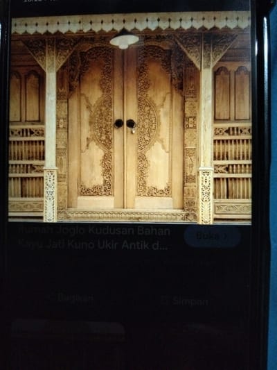 The image displays the ornate wooden doors of a Javanese Joglo house, likely located in Kualaraja, Indonesia. The doors are intricately carved with traditional motifs, featuring decorative panels, a central relief, and two dark, round knobs. To the left and right of the doors are wooden shelves with decorative lattices. Above the doorway, a white, circular lamp is suspended. The architectural style and carvings are characteristic of Javanese culture. The bottom portion of the image contains text overlays indicating "Rumah Joglo Kudusan Bahan Kayu Jati Kuno Ukir Antik d..." and buttons for "Buka >", "Bagikan", and "Simpan". The scene appears to be a digital representation of the house's facade, possibly from a website or application.