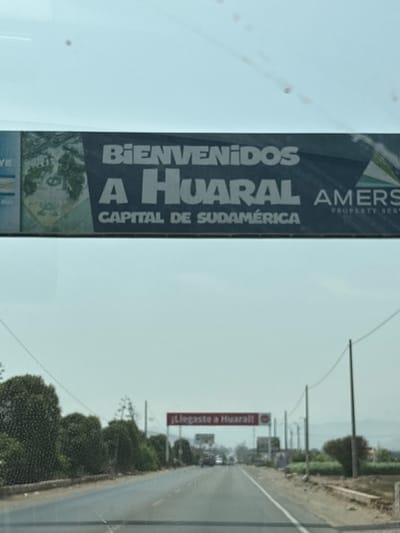 The image shows a roadside view in Aucallama, Peru. A sign welcomes visitors to Huaral, identifying it as the capital of South America. Another sign further down the road displays "Llegaste a Huaral." Utility poles line the road, and dense greenery is visible on either side. The photograph appears to have been taken from inside a vehicle, with a portion of the windshield visible in the foreground.