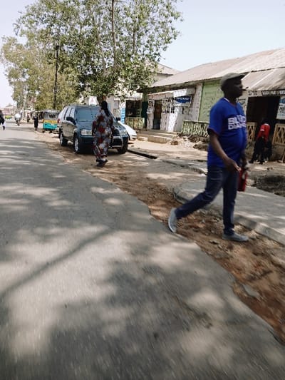 The scene depicts a bustling street in the vicinity of Kungulu School, Zambia, under clear daylight conditions. The foreground consists of an asphalt road displaying tree shadows.
In the foreground right, a dark-complexioned male, blurred due to motion, is walking towards the upper-right of the frame along the edge of the street. He wears a blue t-shirt with "BLUE DIAMOND" text, a dark baseball cap, blue jeans, and grey sneakers. He holds a small red object in his right hand.
In the mid-ground, slightly left of center, a dark-colored SUV, possibly a Honda CR-V, is parked on the street with a chrome bumper guard visible. A dark-complexioned female, wearing a patterned dress, stands next to the SUV, facing away from the viewer.
Further down the street on the left, a light blue and green Bajaj RE (tuk-tuk) is visible, along with a motorcycle or scooter and additional indistinct figures walking.
The right side of the image features a row of single-story commercial buildings with corrugated metal roofs. Their facades are painted in white and green, with storefronts displaying signs, including partially legible text such as "SALOON," "PHARMACY," and "PROVISION STORE." A white, painted railing extends along the front of some of these structures. Several figures are visible near these buildings, one dressed in red.
The environment includes several trees with green foliage casting shadows across the road and a dirt/gravel shoulder separating the road from the buildings on the right. A street light pole is visible on the far left.