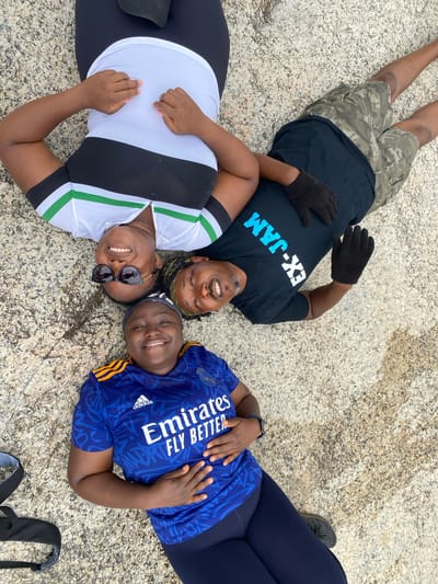 Three adults are lying on a textured stone surface outdoors, arranged in a roughly triangular formation. The person at the top wears a black t-shirt with "EX-JAM" printed on it, camouflage shorts, and gloves. The person on the left wears a white shirt with black sides and green stripes. The person on the right wears a blue soccer jersey with "Emirates FLY BETTER" printed on it and dark leggings. All three are smiling and appear relaxed. The location is Ganawuri, Nigeria.