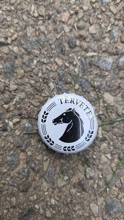 A white metal bottle cap with a black horse head logo and the text "TERVETE" lies on a gravelly, asphalt-like surface. The cap is centrally located and slightly angled. The background consists of small, grey and brown stones mixed with dirt. A few blades of green grass are visible near the cap. The scene is outdoors, likely on a road or path in Cekule, Latvia. No people are present.