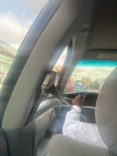 The image depicts the interior of a vehicle, viewed from a passenger's perspective, looking forward towards the driver and the exterior environment in Elimgbu, Nigeria.

**Interior:**
The immediate foreground features light grey fabric upholstery of a seat. To the left, a prominent grey B-pillar is visible, equipped with a light grey plastic seatbelt height adjuster through which a black seatbelt strap is threaded, extending towards the lower right. The vehicle's dashboard, grey in color, is visible beyond the B-pillar and to the right. A steering wheel is visible, with a gauge cluster partially obscured behind it. A light grey headliner and a matching sun visor are visible at the top right. A grey door panel, likely the driver's, is visible, showing a cluster of window control buttons.

**People:**
The right arm and hand of a person with dark skin are visible, firmly gripping the steering wheel. The individual is wearing a white long-sleeved shirt with a visible cuff. The rest of the person's body and face are not visible.

**Exterior Environment:**
Through the vehicle's windshield, an overcast sky is visible. A street scene unfolds, showing multiple vehicles in various states of focus, suggesting traffic. A blue vehicle is centrally located in the mid-distance, and a red vehicle is partially visible to its left. Other indistinct vehicles and structures line the road. To the far left, beyond the vehicle's open window, a building with light-colored or green patterned siding is discernible. Utility poles are faintly visible in the distant background.