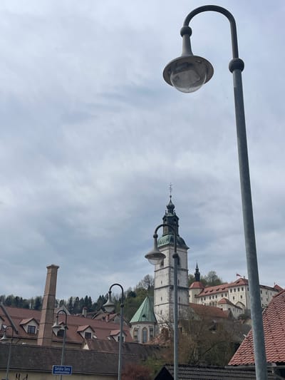 The image shows a street scene in Škofja Loka, Slovenia, with architectural features and urban infrastructure. A prominent lamppost stands in the foreground, partially obscuring the view. In the background is a cityscape, including a tall, white church tower with a spire. Other buildings are visible, displaying a variety of architectural styles and roof types, some with red tiles. A tall chimney is also discernible. The scene takes place under a cloudy sky. A street sign "Selška Sora" is visible. No people are visible in the frame.