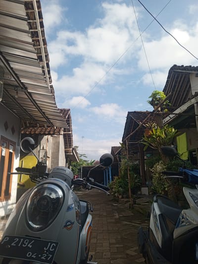 The image depicts a narrow alleyway flanked by houses with traditional tiled roofs. The sky is a bright blue with scattered white clouds, suggesting it is daytime and the weather is fair. Two motorcycles are parked in the foreground, partially obscuring the view of the alley. The motorcycle on the left is white with orange and blue accents and a license plate that reads "P 2194 JC 04.27". The motorcycle on the right is partially visible and appears to be blue and white, with "HONDA" branding.

The houses on either side of the alley have windows and doors visible, and the walls appear to be made of concrete. Lush green plants and vegetation are abundant, growing in pots and trailing from the structures, adding to the tropical feel of the setting. Some plants are also seen growing on the roofs and balconies. A birdcage with a bird inside is hanging from the eaves of the left house. Power lines are stretched across the sky. The overall impression is a glimpse into a residential area in what appears to be a warm climate. No people are visible in the frame. The scene suggests a quiet, ordinary day in this Indonesian locale.