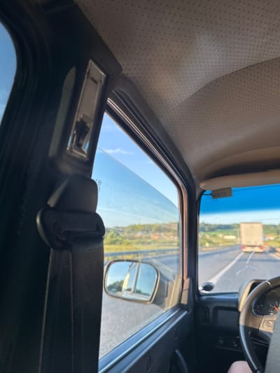 A first-person perspective shot from inside a vehicle driving on a highway during daytime. The view is through the driver's side window, showing the road ahead and to the right, with greenery and a few structures in the distance. A white truck is visible on the highway ahead, driving in the same direction. The interior of the car is visible, including the ceiling lining, a part of the dashboard, the steering wheel, and a seatbelt mechanism. The sun appears to be shining brightly, casting light on the scene. The image does not provide specific details indicating the location is Verkh-Sosnova, Russia.