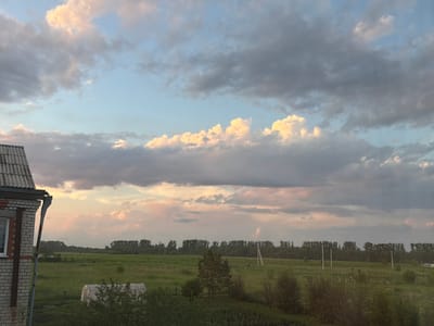The image shows a rural landscape at dusk in Leninsk-Kuznetskiy, Russia. In the foreground, a brick building with a grey roof is partially visible on the left. Beyond the building is a green field dotted with various plants and trees, including a small greenhouse-like structure. In the middle ground, a dense line of trees forms the horizon. The sky is filled with dramatic clouds, some illuminated by the setting sun with hues of pink and orange, while others are dark and grey. Several utility poles are scattered across the field. There are no people visible in the image. The overall scene depicts a tranquil evening in a rural setting.
