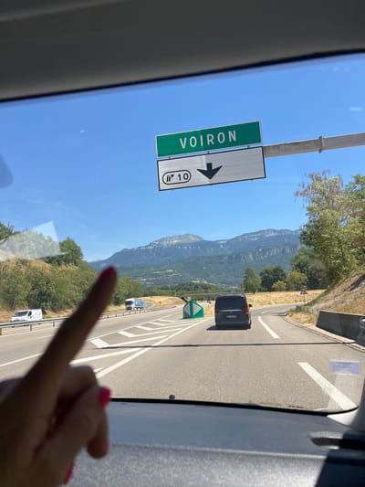 The image is taken from inside a vehicle, looking through the windshield. A person's hand with bright pink fingernails is visible in the foreground, pointing towards the road ahead. A green road sign displaying "VOIRON" in white letters is positioned above a white directional sign with a downward arrow and the number "10" inside a circle. The road ahead is a highway with multiple lanes, and several vehicles are present, including a dark van, a white van, and a truck. The landscape features rolling hills covered with vegetation and a mountain range in the background under a clear blue sky. The scene appears to be on a sunny day. Based on the "VOIRON" sign, the location is likely on a road leading to or from the city of Voiron, France. Given the context of the highway and the scenery, the specific area within or near Apprieu, France is consistent with this type of mountainous terrain and major road infrastructure.