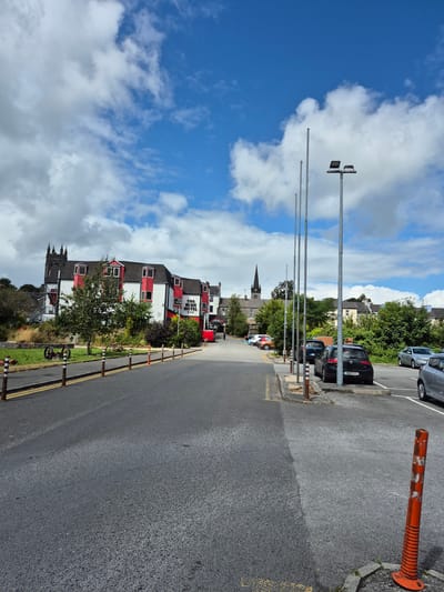 A street scene in Central Park, Ireland, features a road leading towards a building complex with a prominent red and white facade. The building is labeled "THE BUSH HOTEL." To the left of the hotel, a church spire rises against a bright blue sky dotted with white clouds. The road is lined with orange and white bollards. On the right side of the road, a parking lot is visible with several cars parked. One car, a black Volkswagen Golf, has the license plate "141-LH-4103." A red bus is parked in front of the hotel. In the foreground, an orange traffic cone stands on the pavement. The environment appears to be a mix of urban and green spaces, with trees and grass bordering the road and parking area.