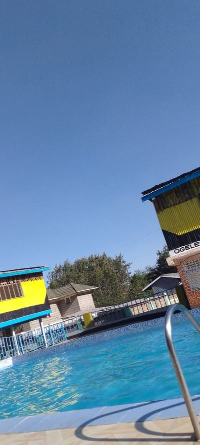 A bright, sunny day reveals a swimming pool surrounded by buildings. The pool's water is a clear blue, and a metal ladder is partially visible. The buildings are characterized by yellow and blue colors, with one structure displaying a sign. A line of trees is visible in the background, partially obscured by the buildings. The clear, blue sky takes up most of the frame.